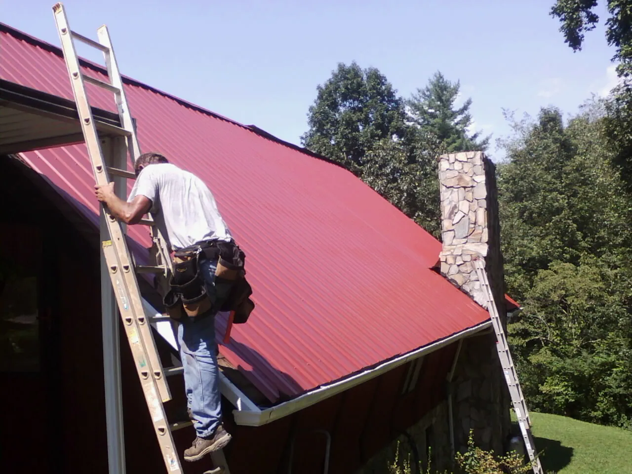Roofer on a ladder beside a red metal roof and stone chimney — Soffit & Fascia Repair by Roofing Brothers in Bulverde